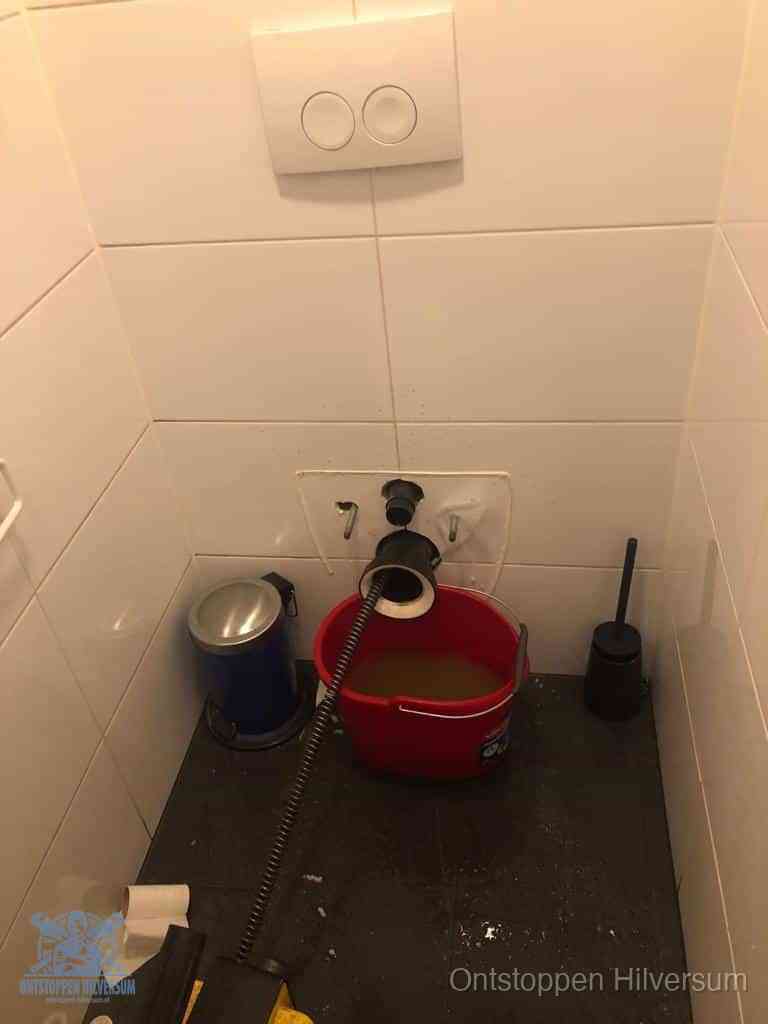 Een ontstopte wand-wc met verwijderde toiletpot waar een ontstoppingsveer in de wanduitgang verdwijnt, het achterhalen waar de verstopping zit in Hilversum