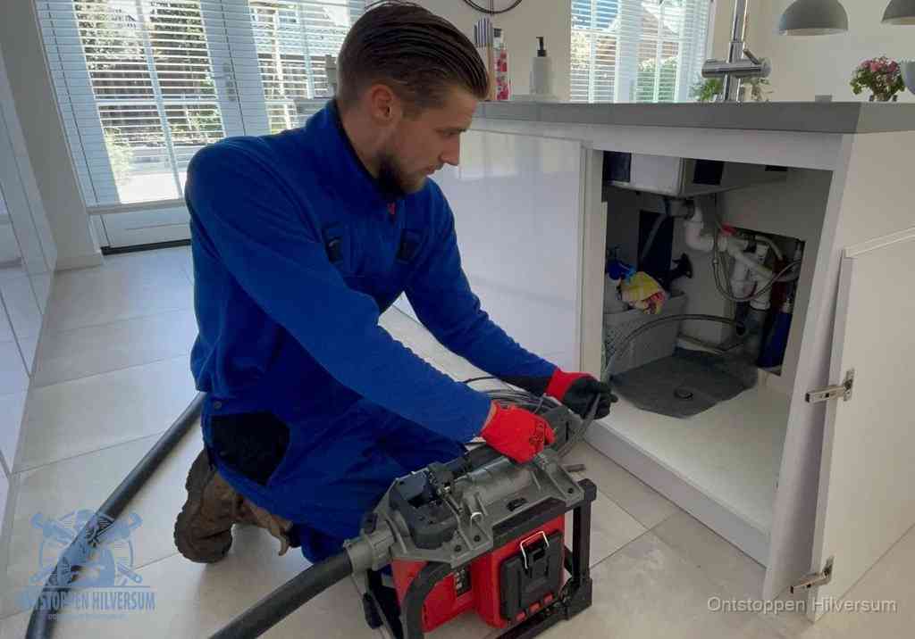 Een monteur voert een professionele elektrische ontstoppingsmachine in de afvoer onder de spoelbak tijdens het gootsteen ontstoppen in Hilversum